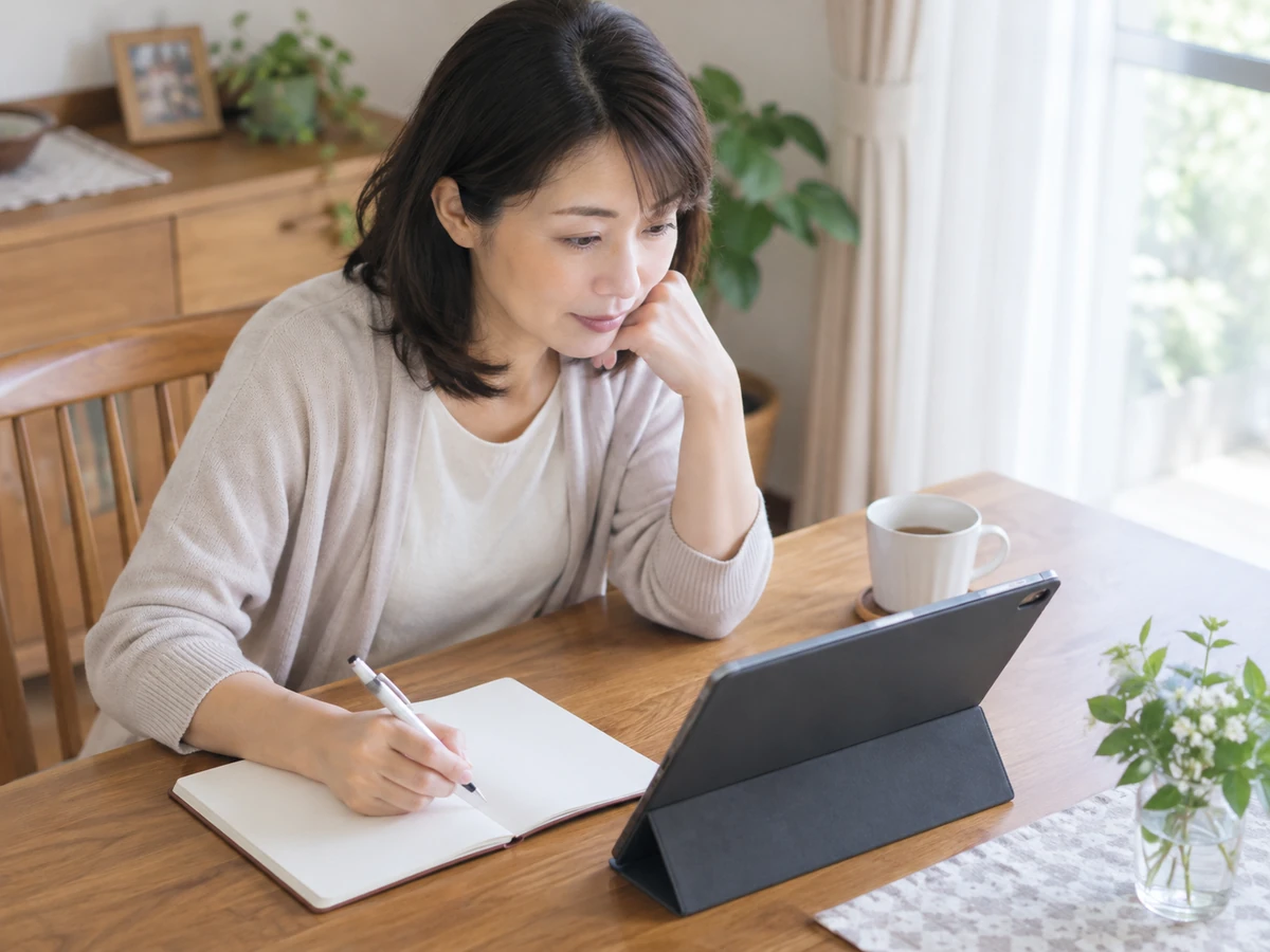 自宅でタブレットを確認しながら、ノートに書き留める女性