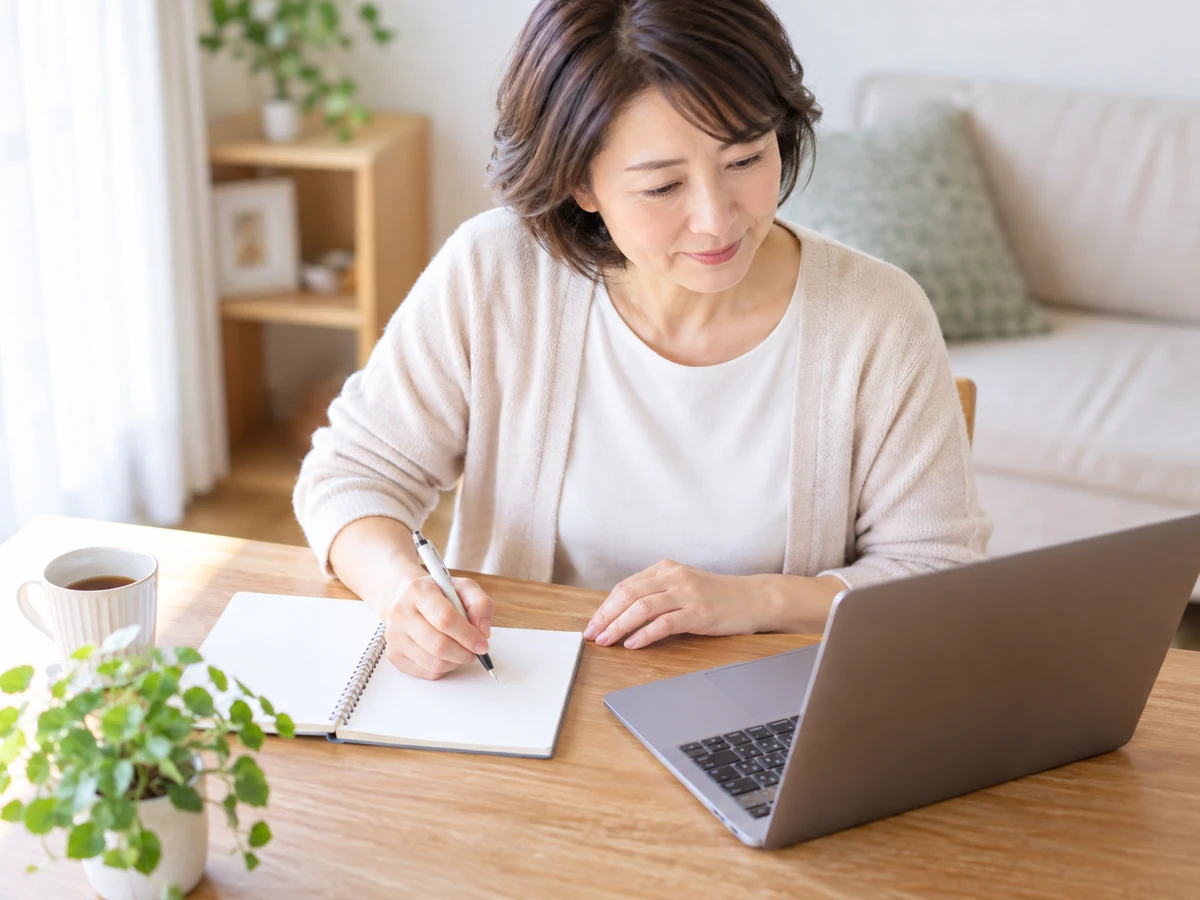 自宅でノートパソコンを見ながら、女性がノートに要点を書き留めている。