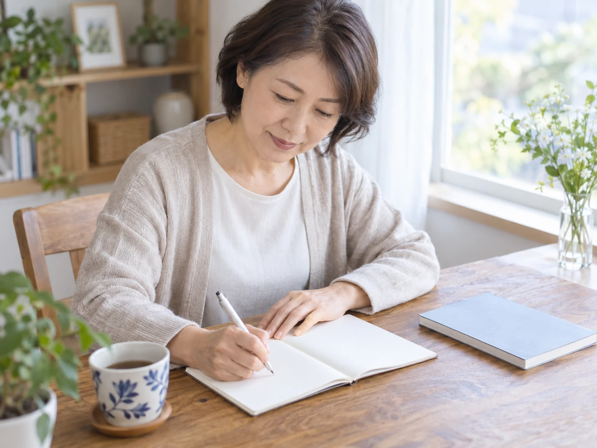 自宅の窓辺でノートに確認したいことを書き留める女性。