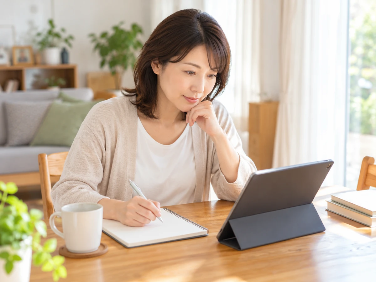 自宅でタブレットを見ながらノートに情報を整理する女性。